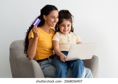 Happy Young Mom And Little Daughter Shopping Online With Laptop Computer And Credit Card, Smiling Millennial Mother And Female Child Purchasing From Internet While Relaxing In Armchair At Home