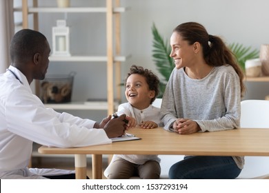 Happy Young Mom And Little Preschooler Son Visiting African American Male Family Doctor Consult On Health Problems, Smiling Mother And Boy Child Talk With Positive Pediatrician In Hospital Or Clinic