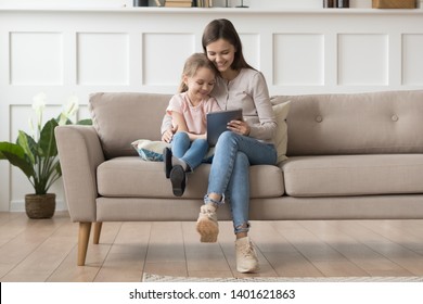 Happy Young Mom Or Nanny Sit On Couch Relax With Little Daughter Watch Cartoon On Tablet Together, Smiling Mother And Preschooler Girl Child Have Fun Using Playing On Pad Resting At Home