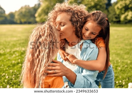Happy young mom sitting in a meadow in the park with her two beloved daughters, hugging and kissing them, having fun and playing on a warm sunny weekend.