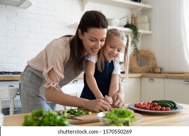 Happy young mom teaching preschool daughter holding knife cut fresh vegetables in kitchen interior, cute little child girl learning cooking prepare salad healthy food with mum enjoy family lifestyle