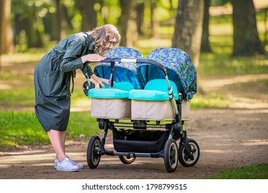 Happy young mom walks in the park in the summer with a stroller for twins, smiling.