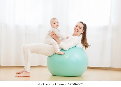 Happy young mother with baby wearing white sport clothes doing gymnastics with Exercise ball and having fun at light domestic home interior.