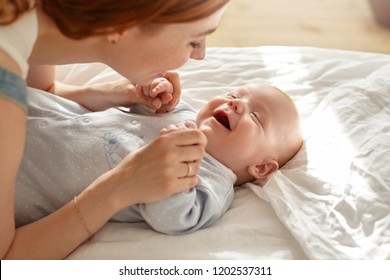 Happy young mother bonding with her toddler son in blue sleeping suit. Sweet portrait of adorable baby lying in bedroom with his mom talking or singing to him. Innocence, togetherness and family