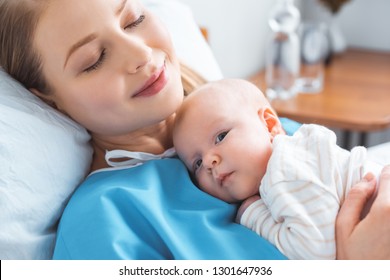 happy young mother with closed eyes lying with adorable newborn baby on chest in hospital room
