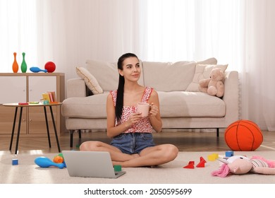 Happy Young Mother With Cup Of Coffee And Laptop Resting In Messy Living Room