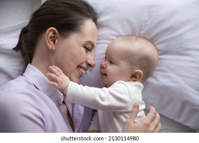 Happy Young Mother And Cute Cheerful Baby Relaxing In Bedroom Together. New Mom Lying On Bed With Pale Linen, Cuddling, Hugging Adorable Infant, Kid Touching Mum Face. Top View, Close Up