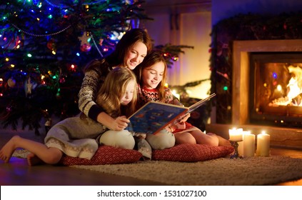 Happy Young Mother And Her Daughters Reading A Story Book Together By A Fireplace In A Cozy Dark Living Room On Christmas Eve. Celebrating Xmas At Home. Winter Evening With Family And Kids.