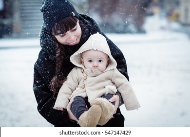 Happy Young Mother Holding Her Newborn Baby In The Cute Sheep Coat With Hood. Mother And New Born Boy Having Good Time In The Park In Winter. Baby Boy Is Looking On The Camera. Color Toned Image.