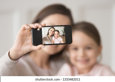 Happy Young Mother Holding Phone Taking Selfie On Cellphone Embracing Child Daughter, Smiling Mom And Kid Girl Looking At Smartphone Camera Making Pic Photo Together, Focus On Mobile Screen Portrait