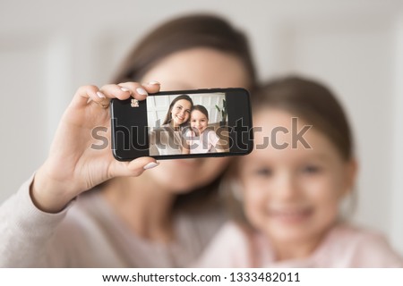 Happy young mother holding phone taking selfie on cellphone embracing child daughter, smiling mom and kid girl looking at smartphone camera making pic photo together, focus on mobile screen portrait