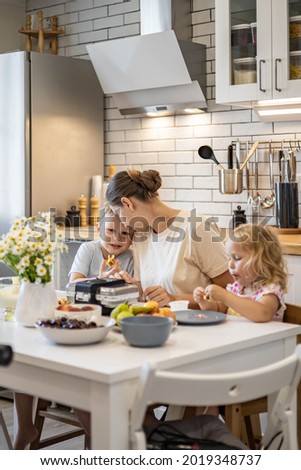 Happy young mother hugging little kids feeling love and tenderness sitting table with fresh homemade summer dessert waffles and fruits. Family weekend breakfast at home kitchen with positive emotion