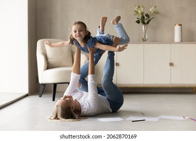 Happy Young Mother Lifting In Air Laughing Cute Little Kid Daughter, Lying On Warm Heated Wooden Floor In Living Room. Joyful Two Female Generations Family Having Fun, Entertaining Togetehr At Home.