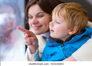 Happy Young Mother And Little Toddler Boy Looking Out Through Window Of A Train, While It Moving. Family, Woman And Funny Cute Kid Son Traveling Together And Making Winter Vacation. Travel Concept.