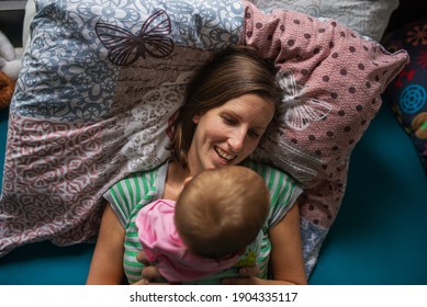 Happy Young Mother Lying On The Bed With Her Baby Girl On Top Of Her In A Genuine Moment Of Joy.