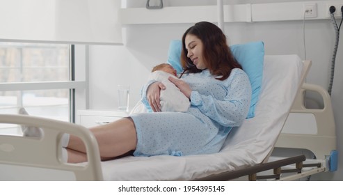 Happy Young Mother With Newborn Baby In Hospital Bed. Portrait Of Smiling Woman After Labor Resting In Bed And Holding Newborn Son Or Daughter In Hospital Ward