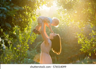 Fröhliche junge Mutter, die mit ihrem kleinen Sohn spielt und sich bei sonnigem Frühling oder Sommertag amüsiert. Schönes Sonnenuntergang im Apfelgarten oder im Park. Fröhliches Familienkonzept