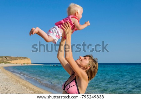 A happy young mother raised her baby girl. In the background there is a sea and a sandy beach. Western Crimea.
