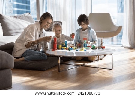 Happy young mother relax at home with little kids, help build castle from colorful wooden blocks, sit at table in living room, develops children, enjoy creative games with daughter and son on pastime