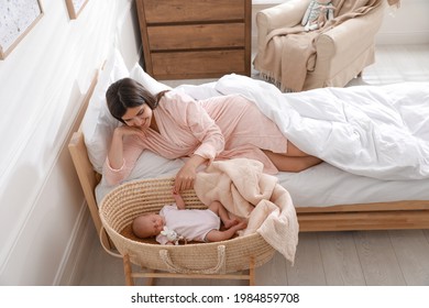 Happy young mother resting near cradle with sleeping newborn baby at home - Shutterstock ID 1984859708