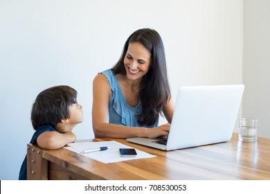 Happy Young Mother Talking To Son While Working On Laptop At Home. Cute Boy Looking Mom While Typing On Computer. Smiling Woman And Child In Conversation While Using Laptop.