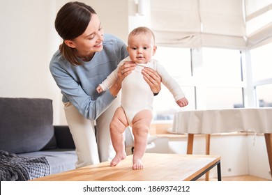 Happy young mother watching her newborn baby son taking first steps at home at the kitchen. Happy childhood and motherhood concept. Stock photo
