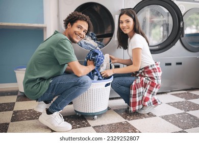 Happy Young Multicultural Couple Doing Laundry Together Loading Washer Machine At Laundromat Room. Guy Washing His Clothes And Posing Smiling At Camera. Public Laundrette Service