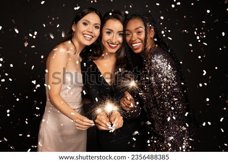 Happy young multicultural girlfriends in nice outfits gesturing posing smiling on black background, holding bengal lights, enjoying falling confetti, celebrating Christmas or New Year party