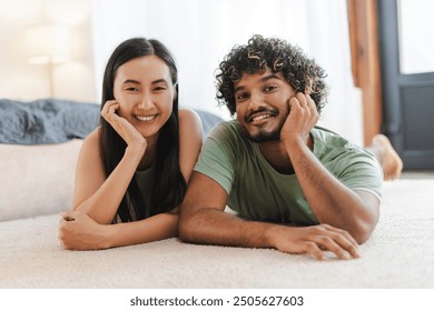 Happy young multi-ethnic couple, smiling Asian woman and Indian man relaxing on carpet at home looking at camera. Relationship concept - Powered by Shutterstock