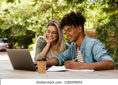 Happy young multiethnic couple spending time together at the park, studying while sitting at the table with laptop