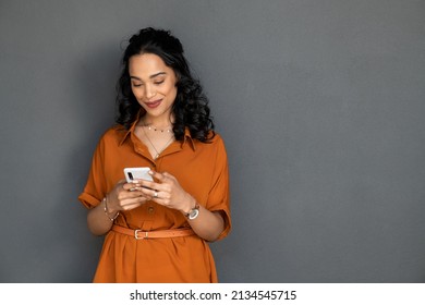 Happy young multiethnic woman writing message on smartphone isolated against grey wall. Beautiful smiling hispanic woman checking mail on mobile phone. Cheerful latin girl chatting by cellphone.