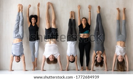Happy young multiracial women in activewear standing near wall with raised arms while sportive mixed race people performing handstands, having fun together before intensive workout in gym indoors.