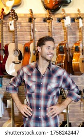 Happy Young Music Store Owner With Hands On Hips Standing In Front Of Guitars