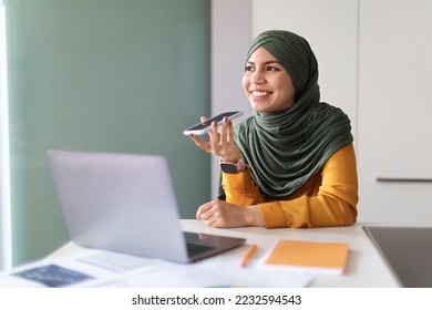 Happy Young Muslim Woman In Hijab Recording Voice Message On Smartphone In Office, Smiling Islamic Female Using Virtual Assistant On Phone, Enjoying Smart Modern Technologies, Copy Space - Powered by Shutterstock