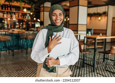 Happy young Muslim woman smiling at the camera while standing in a cafe. Portrait of a young freelancer with a hijab holding a laptop in a coffee shop. - Powered by Shutterstock