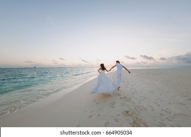 Happy young newly married couple woman in white dress man in love running, have fun on clean sandy beach   waves of azure sea or ocean, summer vacation at water. Wedding rest, relax honeymoon concept.