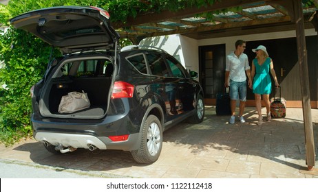 Happy Young Newlyweds Walk Out Of Their Home In The Sunny Suburbs To Pack Their Luggage In The SUV. Girlfriend And Boyfriend Pull Their Travel Bags Behind Them While Walking Towards The Driveway.