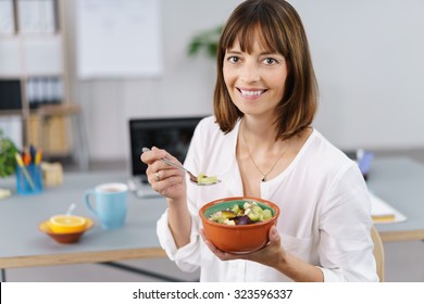 Happy Young Office Worker Enjoying A Healthy Lunch Sitting At A Table At Work Eating A Bowl Of Fresh Fruit Salad And Looking At The Camera With A Vivacious Smile