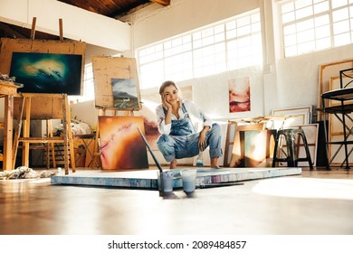 Happy young painter squatting on the floor while working in her art studio. Female artist smiling at the camera and holding a paintbrush. Creative young woman making a new painting on a canvas.
