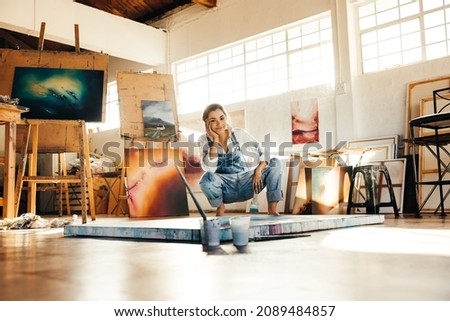 Happy young painter squatting on the floor while working in her art studio. Female artist smiling at the camera and holding a paintbrush. Creative young woman making a new painting on a canvas.