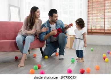 Happy young parents Caucasian father and Asian mother playing ukulele and singing with little kid girl in living room at new house. Full of boxes and colorful balls on floor.