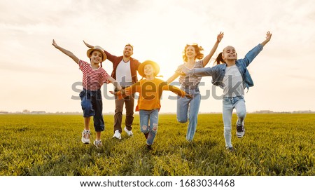 Happy young parents with daughters in casual clothes running with outstretched arms while enjoying time together on green meadow in summer evening
