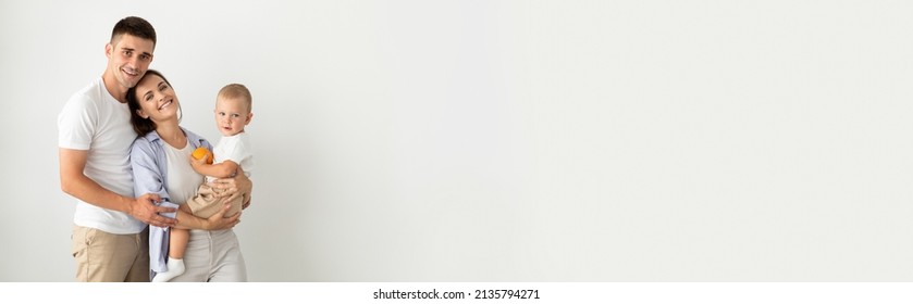 Happy Young Parents Holding Their Cute Toddler Son And Smiling At Camera, Portrait Of Beautiful Cheerful Family Of Three With Little Baby Posing Together Over White Background, Copy Space, Panorama