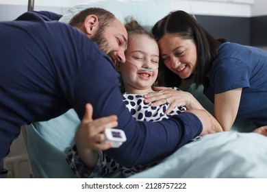 Happy Young Parents Hugging Hospitalized Recovered Little Daughter While In Pediatric Clinic Patient Room. Cheerful Family Laughing And Hugging In Health Pediatric Hospital Room.