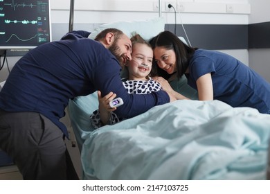 Happy young parents hugging ill hospitalized recovering little girl while in pediatric hospital patient room. Positive joyful family laughing and hugging in health pediatric clinic room.