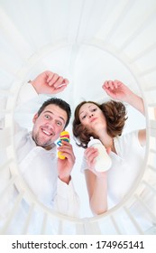 Happy Young Parents Making Funny Faces Looking At Their Baby In A White Round Crib - From Baby Perspective View