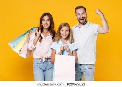 Happy young parents mom dad with child kid daughter teen girl in basic t-shirts hold package bags with purchases doing winner gesture isolated on yellow background studio portrait. Family day concept Foto Stock