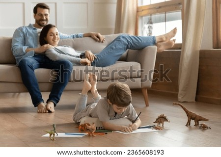 Happy young parents relaxing on couch while adorable little son drawing colorful pencils, playing on warm wooden floor with underfloor heating, family enjoying leisure time on weekend at home