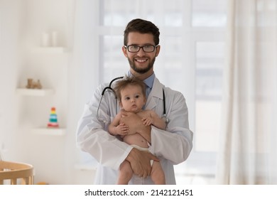 Happy Young Pediatrician Man Wearing White Coat, Holding Baby Patient In Arms, Looking At Camera, Smiling. Kids Doctor With Few Month Child Head Shot Professional Portrait. Infant Health Care