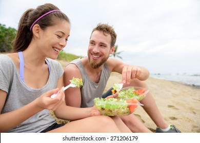 Happy young people eating healthy salad for lunch. Multiracial group having a break on beach snacking on a vegan takeaway meal of green veggies and carrots laughing together. Casual lifestyle.